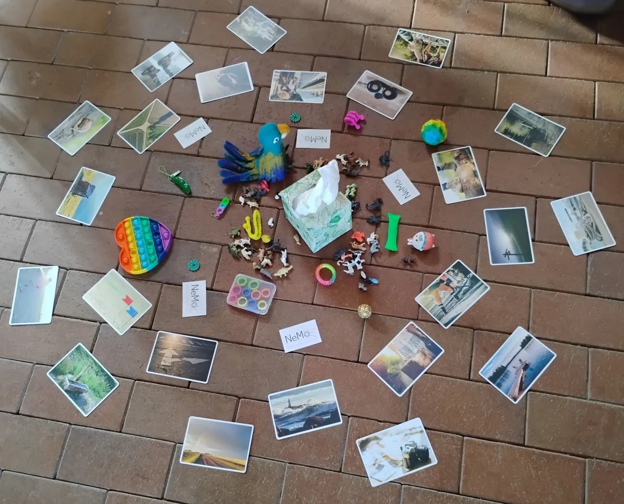 Fotokarten, kleine Figuren und Karten mit der Aufschrift „NeMo“ liegen im Kreis auf einem Fliesenboden