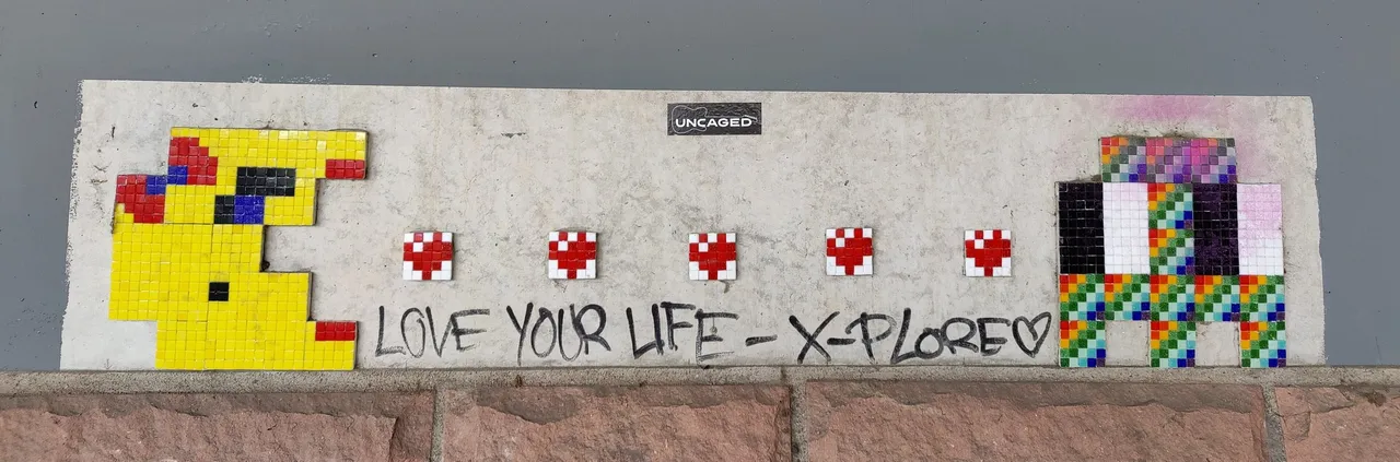 Mosaik an einer Hauswand: eine bunte Figur, Herzen und der Schriftzug „Love your life – X-plore“